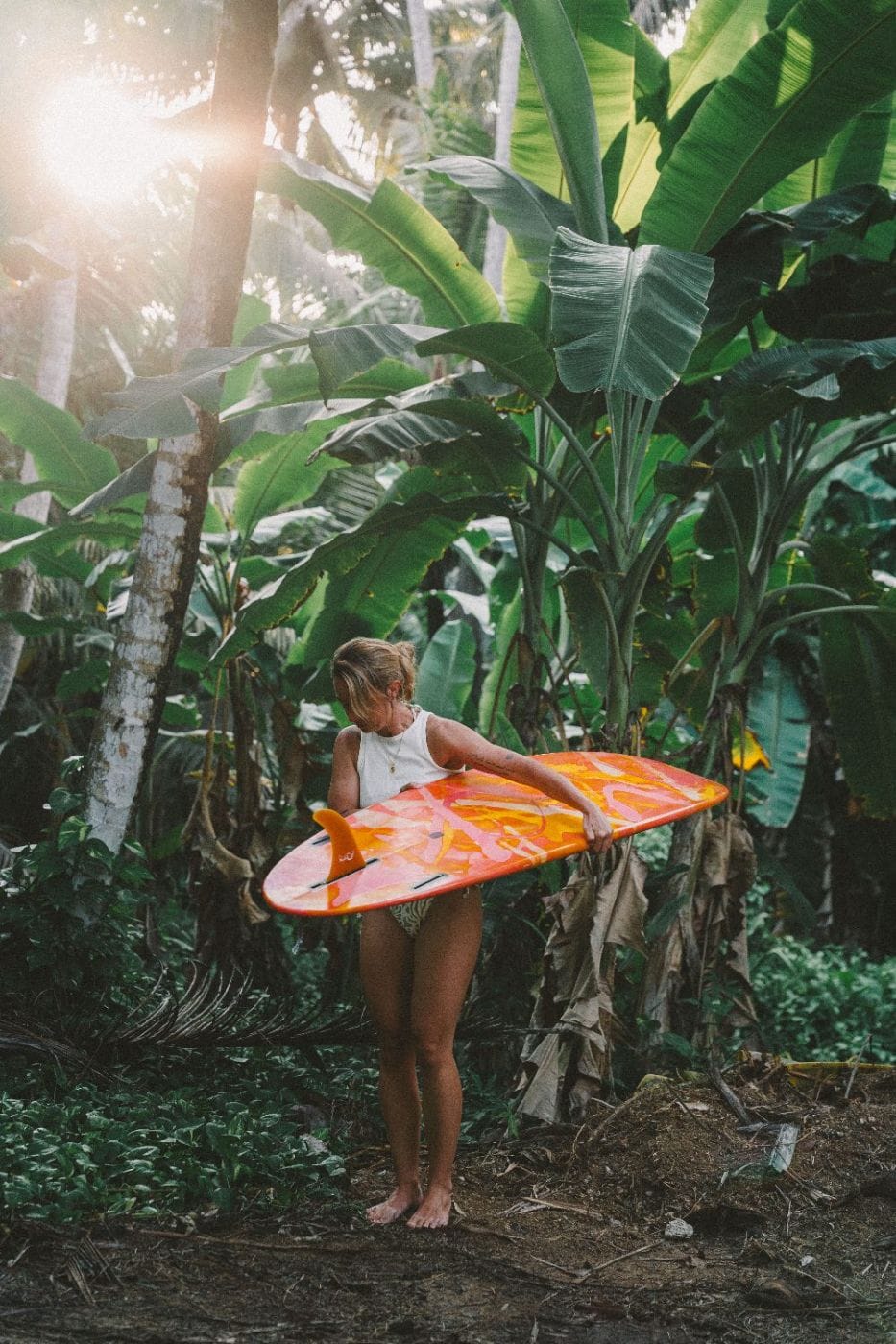 A surfer carrying her board through the banana palms at golden hour in Madiha